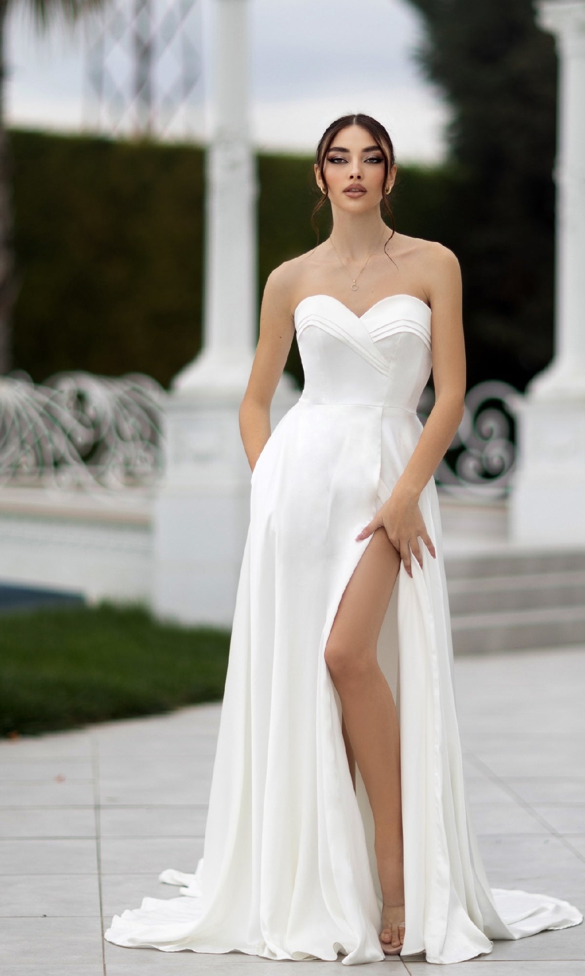 A woman wearing the Off-White Strapless Bridal Gown: Tina Holly TW105, featuring a thigh-high slit and sweetheart neckline, stands outdoors on a tiled surface, posing with one hand holding the dress.